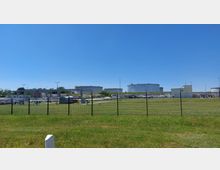 Weitläufiges Industriegelände mit großen, zylindrischen Tanks im Hintergrund, umgeben von Zäunen. Im Vordergrund sind Rasenflächen und Metallrohre sichtbar, die das Gelände durchziehen.