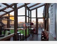 Innenraum eines Wintergartens mit Holzkonstruktion und großen Glasfenstern, die einen Blick auf eine Terrasse mit Sitzgelegenheiten und Lichterketten bieten. Im Vordergrund sind Tische, Stühle, Barhocker und Kleidungsständer sichtbar, im Hintergrund sieht man eine städtische Dachlandschaft.