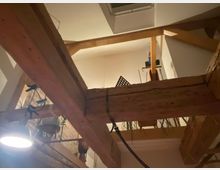 Ein offener Innenraum mit sichtbaren Holzträgern und einer Zwischendecke. Im Hintergrund sind eine Regalstruktur, ein Notenständer und hängende Pflanzen sichtbar. Ein funktionales Deckenlicht leuchtet den Raum aus.