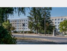 Mehrstöckiges Wohngebäude mit heller Fassade und zahlreichen rechteckigen Fenstern, umgeben von Bäumen und Grünflächen. Im Vordergrund befinden sich eine Straße, ein Gehweg und ein Verkehrsschild.