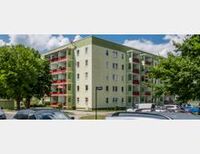 Wohnblock mit vier Etagen, grün gestrichener Fassade und Balkonen mit roten Brüstungen, umgeben von Bäumen und geparkten Autos. Der Himmel ist klar mit einigen Wolken.