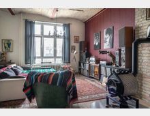 Wohnzimmer mit gemütlicher Einrichtung und Retro-Elementen. Zu sehen sind ein weißes Sofa mit Decken, ein Sessel, ein Teppich mit orientalischem Muster, eine wandhohe Fensterfront mit Vorhängen, eine rote Wand mit Bildern, ein Regal mit Schallplatten und Lautsprechern sowie ein Ofen vor einer Ziegelwand.