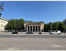 Neoklassizistisches Gebäude mit Säulenvorhalle, umgeben von Bäumen; davor eine gepflasterte Straße mit parkenden Autos und einer Laterne. Im Hintergrund wehen deutsche und andere Nationalflaggen.
