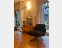 Modernes Wohnzimmer mit Holzparkettboden, einem schwarzen Sessel und einem Holz-Sideboard mit einem Spiegel darauf. Große Fenster mit weißen Vorhängen bieten Blick auf einen bewaldeten Garten.