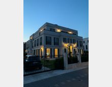 Ein modernes, zweistöckiges Wohngebäude im klassischen Stil mit weißen Fassaden und schwarzen Fensterläden, beleuchtet in der Abenddämmerung. Ein gepflegter Garten mit Zaun und geparktem Auto im Vordergrund.