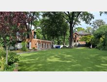 Grüne Gartenanlage mit zwei roten Backsteingebäuden; im Vordergrund eine große Rasenfläche, umgeben von Bäumen und Sträuchern. Eines der Gebäude hat ein modernes, eingeschossiges Design mit vielen Fenstern, das andere ist ein zweigeschossiges Wohnhaus mit Satteldach.