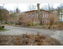 Backsteinbau mit großen Fenstern, umgeben von kahlen Bäumen und Sträuchern in einer leicht vernachlässigten Umgebung. Im Hintergrund sind weitere Gebäude und ein Kirchturm mit grünem Spitzdach zu sehen.
