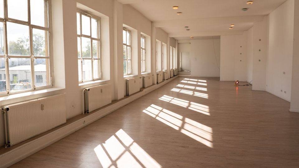 Langer, leerer Innenraum mit weißen Wänden, hellem Holz- bzw. Laminatboden und einer Reihe großer Sprossenfenster entlang der linken Seite. Unter den Fenstern stehen Heizkörper, und das Tageslicht wirft rechteckige Schattenmuster auf den Boden.