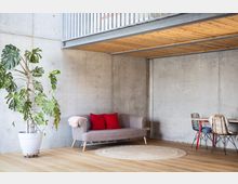 Modernes Wohnzimmer mit Sichtbetonwänden, einem Holzboden und einer Decke aus Holzlatten. Im Raum stehen ein graues Sofa mit roten Kissen, ein großer Topfpflanze sowie ein Essbereich mit Tisch und geflochtenen Stühlen.