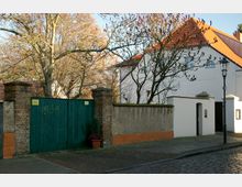 Ein gepflasterter Gehweg und eine Straße führen an einem Grundstück mit einer hohen Mauer aus verputztem Beton und Backstein entlang. Hinter einem großen grünen Holztor und kahlen Bäumen ist ein weißes Gebäude mit roten Ziegeldächern zu sehen.
