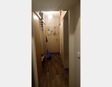 Ein schmaler Flur mit Holzfußboden, Wandschränken auf beiden Seiten und einer Tür am Ende, auf der Zettel und Magnete befestigt sind. Die Decke ist mit einer einzelnen, einfachen Lampe beleuchtet.