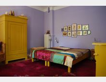 Schlafzimmer mit lavendelfarbenen Wänden, rotem Teppichboden und einem Bett mit bunter Patchworkdecke. Links steht ein großer gelber Kleiderschrank, an der Wand hängen mehrere gerahmte Bilder, daneben sind ein Heizkörper und ein kleiner Nachttisch zu sehen.
