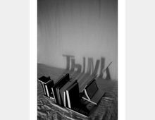 Schwarz-weiß-Fotografie eines Bücherstapels, dessen Schatten an eine Wand das Wort "THINK" projiziert. Die Szene zeigt Bücher auf einem Tisch mit einer einfachen, strukturlosen Hintergrundwand.