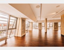 Innenraum eines modernen Gebäudes mit großzügigen Fenstern, die eine schräg geneigte Glasfassade bilden und den Blick auf eine städtische Umgebung freigeben. Der Raum hat einen dunklen Holzboden, helle Wände, eine Säule und integrierte Beleuchtung in der Decke.