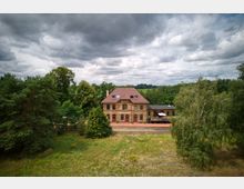 Historisches Bahnhofsgebäude aus gelbem Backstein mit rotem Satteldach, gelegen in einer ländlichen Gegend mit Bäumen und Wiesen im Hintergrund. Vor dem Gebäude verlaufen Bahngleise, daneben steht ein Güterwaggon auf einem Nebengleis.