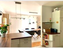 Moderne helle Wohnküche mit offenem Übergang zum Essbereich. Zu sehen sind eine Küchenzeile mit Holzar-beitsplatte und einem Regal mit Büchern, ein Holztisch mit schwarzen Stühlen sowie eine helle Wand mit Kaminelement und großer Fensterfront.