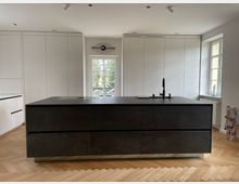 Moderne Küche mit einer schwarzen Kücheninsel in der Mitte, ausgestattet mit einem Wasserhahn und integriertem Kochfeld. Im Hintergrund weiße, glatte Einbauschränke, ein Fenster mit Blick ins Freie und ein Fischaugen-Wandaccessoire; heller Holzparkettboden ergänzt das minimalistische Design.