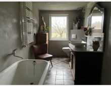 Helles Badezimmer mit freistehender Badewanne, einem Waschtisch mit Aufsatzwaschbecken und Spiegel, sowie einem Fenster, das auf eine grüne Landschaft blickt. Der Raum verfügt über graue Wände, gemusterte Bodenfliesen und dekorative Elemente wie Pflanzen und eine Vintage-Holzkommode.