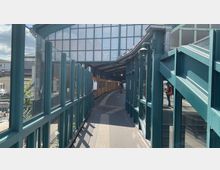 Überdachte U-Bahn-Station mit gelbem Zug, großen Glasfenstern und grünem Metallrahmen. Im Vordergrund ist ein schmaler Gehweg, im Hintergrund sind Personen und der Eingang zur Station sichtbar.