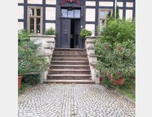 Fachwerkhaus mit hellem Putz und dunklen Holzbalken, zentraler Eingangstreppe aus Stein mit dekorativen Geländern, umgeben von grüner Bepflanzung und blühenden Zierpflanzen in Töpfen auf einem gepflasterten Weg.