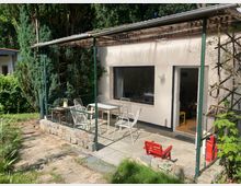 Überdachte Terrasse an einem eingeschossigen Haus mit weißer Fassade, großem Fenster und offener Tür. Auf dem gepflasterten Bereich stehen mehrere Metallstühle und Tische, umgeben von niedriger Steinmauer und dichtem Baumbestand im Garten.