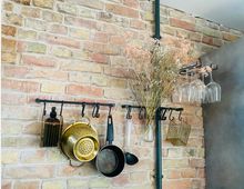 Ziegelmauer mit schwarzer Metallstange, an der Küchenutensilien wie eine goldene Metallschale, eine Pfanne und Glasflaschen aufgehängt sind; daneben trockene Blumen in einem Glasgefäß und umgedrehte Weingläser in einem Halter.