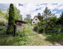 Blick durch ein Drahtzauntor auf ein kleines Einfamilienhaus mit steilem Satteldach am Ende eines schmalen Zufahrtswegs. Der Vorgarten ist dicht bewachsen mit Sträuchern und hohen Bäumen; links steht ein Holzunterstand mit gestapeltem Brennholz.