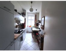 Narrow galley kitchen with white cabinets and a long countertop with sink and stove along the left wall, leading to a small dining table near a large window. The room has light tile flooring, pendant lights, potted plants on the windowsill, and a side shelf with glassware and a wall-mounted board on the right.