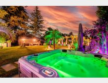 Gartenlandschaft bei Sonnenuntergang mit beleuchtetem Whirlpool im Vordergrund. Der Garten ist mit Palmen und anderen Pflanzen geschmückt, farbige Lichter und Sitzbereiche schaffen eine lebhafte, tropische Atmosphäre.