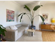 Bright living room with white walls and light wood floors, featuring a light gray sectional sofa on a textured rug and a small marble-top coffee table. A tall potted plant with large leaves stands near a wooden sideboard with a yellow lamp, and colorful framed abstract artwork hangs on the walls beneath a modern white pendant light.
