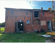 Ziegelsteingebäude auf einem grasbewachsenen Grundstück, mit offenen Toren und kleinen Fenstern. Das Gebäude zeigt altersbedingte Spuren und Renovierungsarbeiten, während im Vordergrund ein Baukran sichtbar ist.