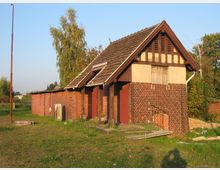 Ein kleines, verfallenes Backsteingebäude mit Fachwerkelementen im Giebel, umgeben von Gras und Bäumen. Das Dach ist mit Dachziegeln gedeckt, und Teile der Fassade wirken beschädigt; im Vordergrund liegen Holzbretter und andere Trümmer auf dem Boden.