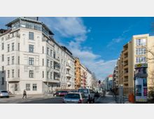 Straßenszene in einem städtischen Wohngebiet mit mehrstöckigen Wohnhäusern im Altbau- und Neubaustil. Links ist ein helles, weißes Eckgebäude mit Balkonen und großen Fenstern, rechts gelbe Fassaden, parkende Autos und ein Gehweg mit Schildern und einem Mülleimer.