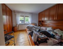 Schlafzimmer mit Holzfußboden, großem Bett und wandhohen Holzschränken an beiden Seiten. In der Mitte ein Fenster mit weißen Gardinen und Heizkörper darunter, daneben einfache Sitzmöbel und ein Korb.