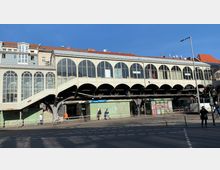 Außenansicht des U-Bahnhofs Görlitzer Bahnhof in Berlin, mit einem erhöhten Gebäude im typischen architektonischen Stil von Hochbahnhöfen, erkennbar an den großen, bogenförmigen Fenstern und einer außenliegenden Treppe. Im Vordergrund befinden sich eine Straße, Gehwege und Menschen.