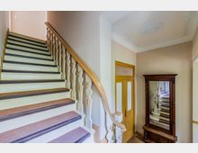 Innenansicht eines Altbau-Treppenhauses mit breiter Holztreppe, hell gestrichenen Stufen, Teppichläufer und geschwungenem Geländer mit Zierelementen. Daneben stehen eine gelbe Innentür mit Glasfeld und ein großer, dunkel gerahmter Spiegel; die Decke hat Stuckornamente.