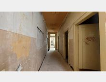 Ein langer, schmaler Flur in einem verlassenen Gebäude mit abgenutzten Wänden, abgeblätterter Farbe und teils freiliegendem Putz. Türen zu mehreren Räumen sind auf der rechten Seite sichtbar, und am Ende des Flurs fällt Licht durch ein Fenster.