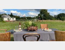 Ein gedeckter Tisch im Freien mit einer grauen Tischdecke, einer Blumenvase, einer Flasche Sekt, Gläsern und Obst. Im Hintergrund eine grüne Wiese, ein Volleyballnetz, ein weißer Anhänger und eine Holzhütte, umgeben von Bäumen.