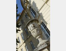 Steinernes Zierelement in Form einer antiken Vase im Vordergrund, vor der Fassade eines historischen Gebäudes mit gelbem Außenputz, hohen Rundbogenfenstern und architektonischen Verzierungen. Schatten von Bäumen fallen auf die Fassade.