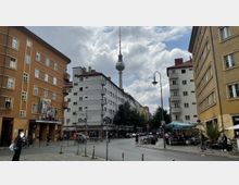 Straßenszene in Berlin mit dem markanten Fernsehturm im Hintergrund, umgeben von mehrstöckigen Wohngebäuden im Stil der Nachkriegsarchitektur. Im Vordergrund sind ein Kino („Babylon“), Straßencafés, Fahrräder und einige Passanten zu sehen.