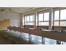 Verlassener, leerer Werkraum mit langen Reihen von Arbeitstischen und montierten Hebeln/Bedienelementen, vor großen Fenstern mit Heizkörpern darunter. Die Decke zeigt abblätternde Farbe, und der Betonboden ist stellenweise mit abgelösten Farbresten bedeckt.