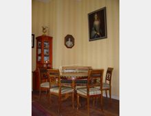 Ein historisch eingerichtetes Zimmer mit einer runden, hölzernen Tischgruppe und vier Stühlen mit gepolsterten Sitzen. An der beigefarben tapezierten Wand hängen zwei Porträtgemälde, und in der linken Ecke steht eine Vitrine mit Glasfront.