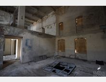 Innenraum eines verlassenen, zweigeschossigen Industrie- oder Lagergebäudes mit rohen Betonpfeilern, bröckelndem Putz und zugemauerten bzw. verbretterten Fensteröffnungen. Auf dem staubigen Boden liegen Metallrahmen und Schutt, in der Mitte befindet sich eine offene Bodenaussparung.