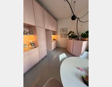 Eine moderne Küche mit rosa Einbauschränken, grauem Fußboden und einem runden weißen Esszimmertisch im Vordergrund. Auf den Schränken stehen Küchenutensilien, Pflanzen und eine Wanddekoration ist sichtbar.