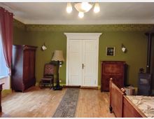 Wohnzimmer im traditionellen Stil mit grünen Wänden und Holzboden. Der Raum enthält antike Möbel wie einen Schrank, eine Kommode, ein Bett und Lampe sowie eine weiße Doppeltür in der Mitte der Wand. Ein Kaminofen steht in der rechten Ecke.