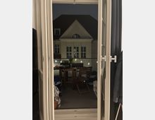 Blick durch eine geöffnete Glastür auf einen beleuchteten Balkon mit einem Holztisch und -stühlen, dekoriert mit Lichterketten. Im Hintergrund ist die Fassade eines Gebäudes mit Satteldach und Sprossenfenstern sichtbar, aufgenommen bei Nacht.