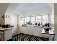 Helle Küche im Retro-Stil mit weißer Einrichtung, schwarz-weiß kariertem Fliesenboden und einem großen Fenster mit Sprossen. Auf der Fensterbank stehen Pflanzen und Küchengeräte, die Arbeitsflächen sind schlicht gehalten.