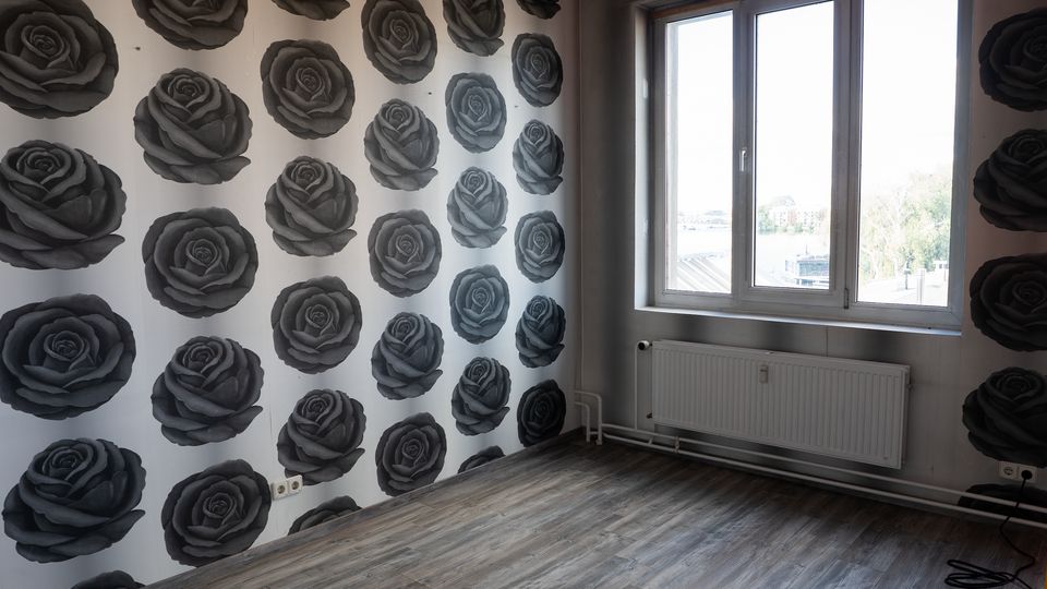 Leerer Innenraum mit grauem Laminatboden und einer Wandtapete mit großen, dunklen Rosenmotiven. Rechts befindet sich ein großes Fenster mit Heizkörper darunter sowie sichtbaren Rohrleitungen entlang der Wand.
