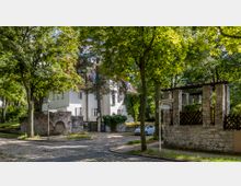 Großer Wohnbereich mit einem weißen, zweistöckigen Gebäude im Hintergrund, umgeben von Bäumen und Grünflächen. Im Vordergrund eine gepflasterte Straße, ein Steinzaun mit Torbogen und Straßenschilder, die auf "Lohengrinstraße" und "Walhallastraße" hinweisen.