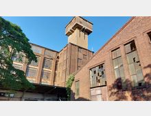 Backstein-Industriegebäude mit großen, teilweise zerbrochenen Fenstern und einem dominanten Wasserturm in der Mitte. Im Vordergrund ein Baum und ein überdachter Bereich, der an die Fassade angrenzt.
